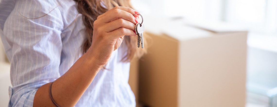 Young woman showing or giving key, posing in new apartment with heap of carton boxes in background. Cropped shot, closeup. Property buying or mortgage concept